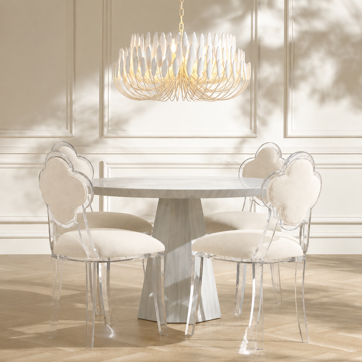A dining room scene with a round white table and four clear acrylic chairs with cream upholstered backs and seats, under a large white leaf-shaped chandelier.