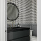 A modern bathroom vanity featuring a glossy black double-drawer cabinet, a black rectangular sink, and a round black-framed mirror. The wall behind the vanity is covered in a black and white geometric patterned tile, with gray wainscoting below and a white toilet visible on the right.