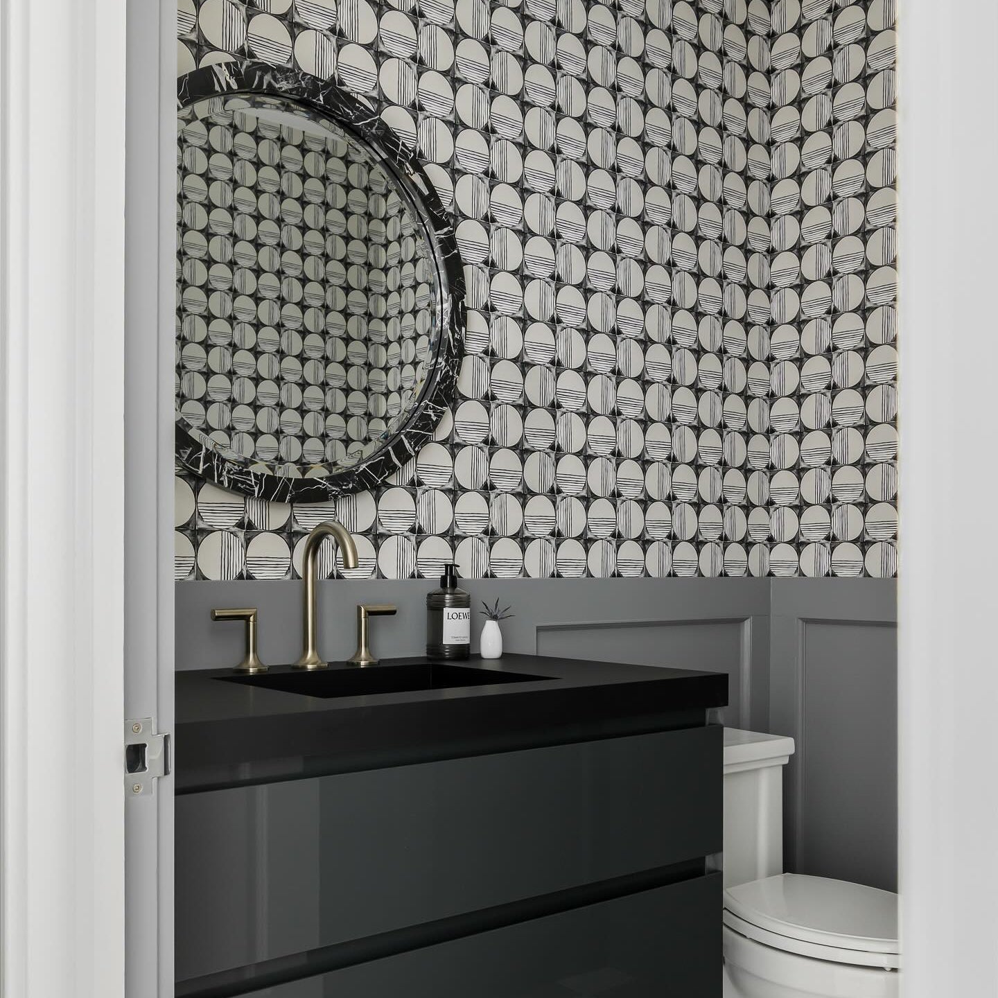 A modern bathroom vanity featuring a glossy black double-drawer cabinet, a black rectangular sink, and a round black-framed mirror. The wall behind the vanity is covered in a black and white geometric patterned tile, with gray wainscoting below and a white toilet visible on the right.