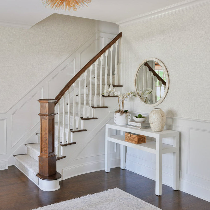 An elegant interior entryway featuring a staircase with a dark wood handrail and white spindles, a light blue console table with decorative items, a round wall mirror, and a textured orange starburst ceiling light.