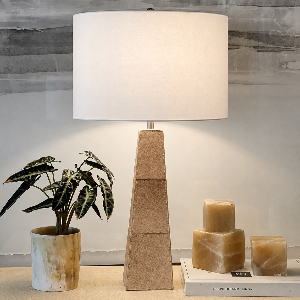 A table lamp with a tapered rectangular beige stone base and a large white drum shade, styled on a tabletop with a potted plant and candles against a light wall.