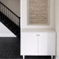 A framed abstract textured wall art piece with beige and brown tones, displayed in a hallway above a white cabinet with gold handles, next to a black staircase.
