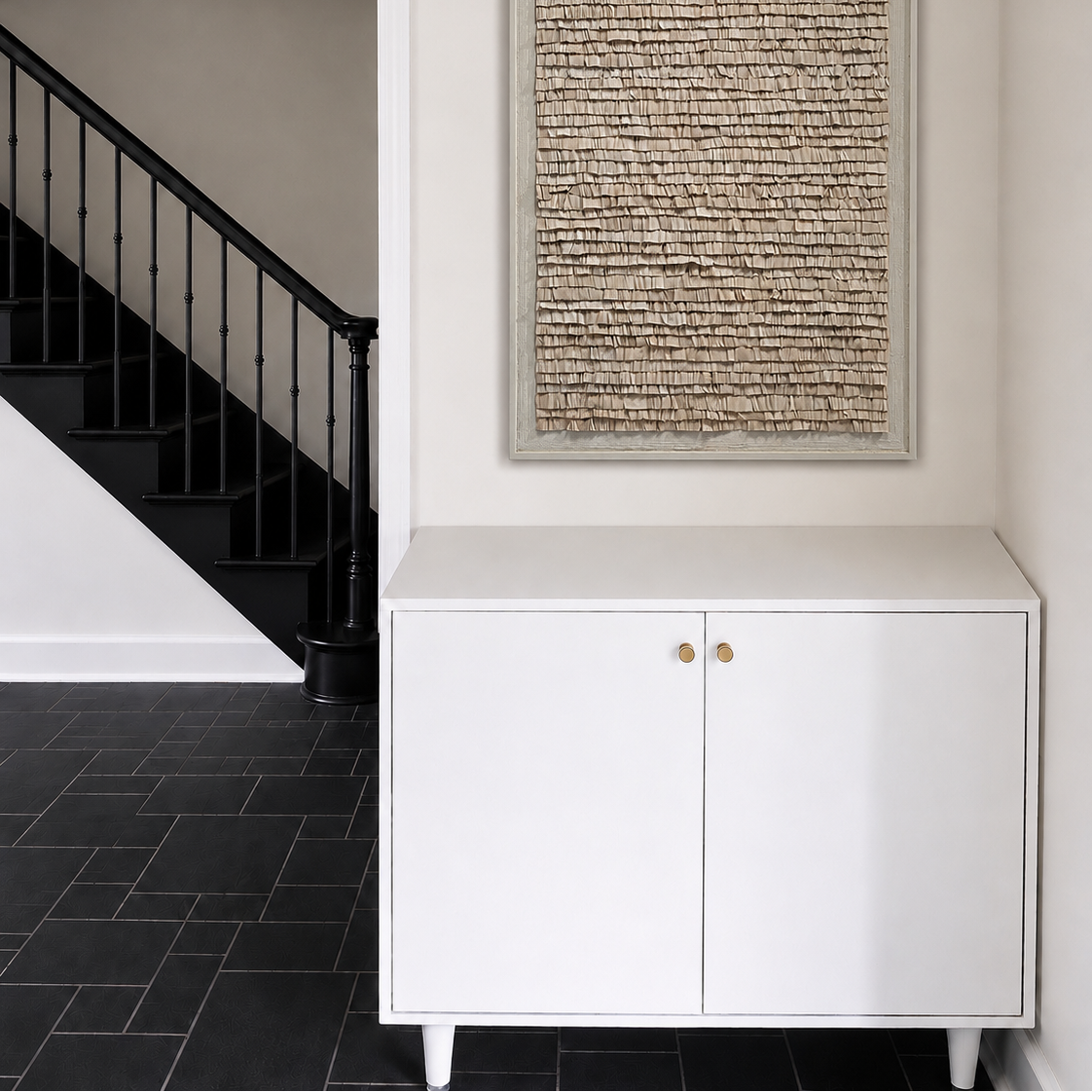 A framed abstract textured wall art piece with beige and brown tones, displayed in a hallway above a white cabinet with gold handles, next to a black staircase.