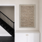 A framed abstract textured wall art piece with beige and brown tones, displayed in a hallway above a white cabinet with gold handles, next to a black staircase.