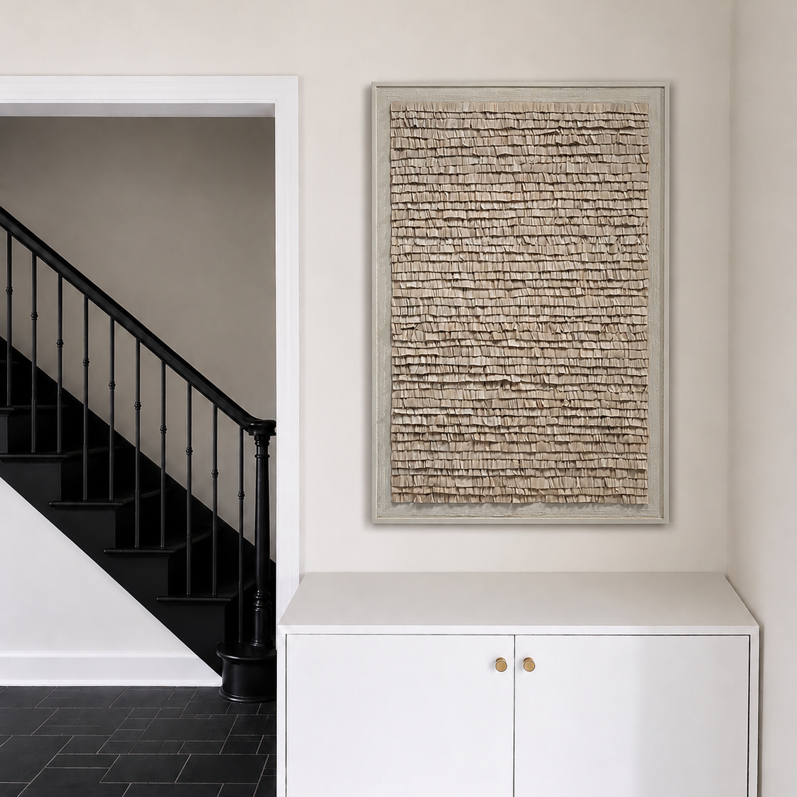 A framed abstract textured wall art piece with beige and brown tones, displayed in a hallway above a white cabinet with gold handles, next to a black staircase.