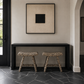 A minimalist interior scene with two leopard-print stools beneath a black console table, in front of a beige wall with a framed black square artwork. A dark doorway and stone floor are visible.
