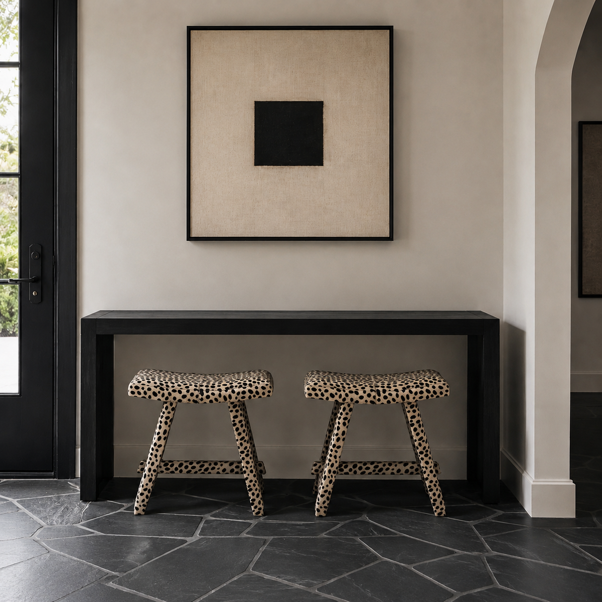 A minimalist interior scene with two leopard-print stools beneath a black console table, in front of a beige wall with a framed black square artwork. A dark doorway and stone floor are visible.