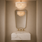 A modern bathroom vanity setup featuring a rectangular white marble sink with gold hardware, a large gold-framed mirror, and a matching gold-framed wall sconce reflecting the light. Above the mirror, a round gold-framed chandelier with a white fringed shade hangs against a warm beige textured wall.