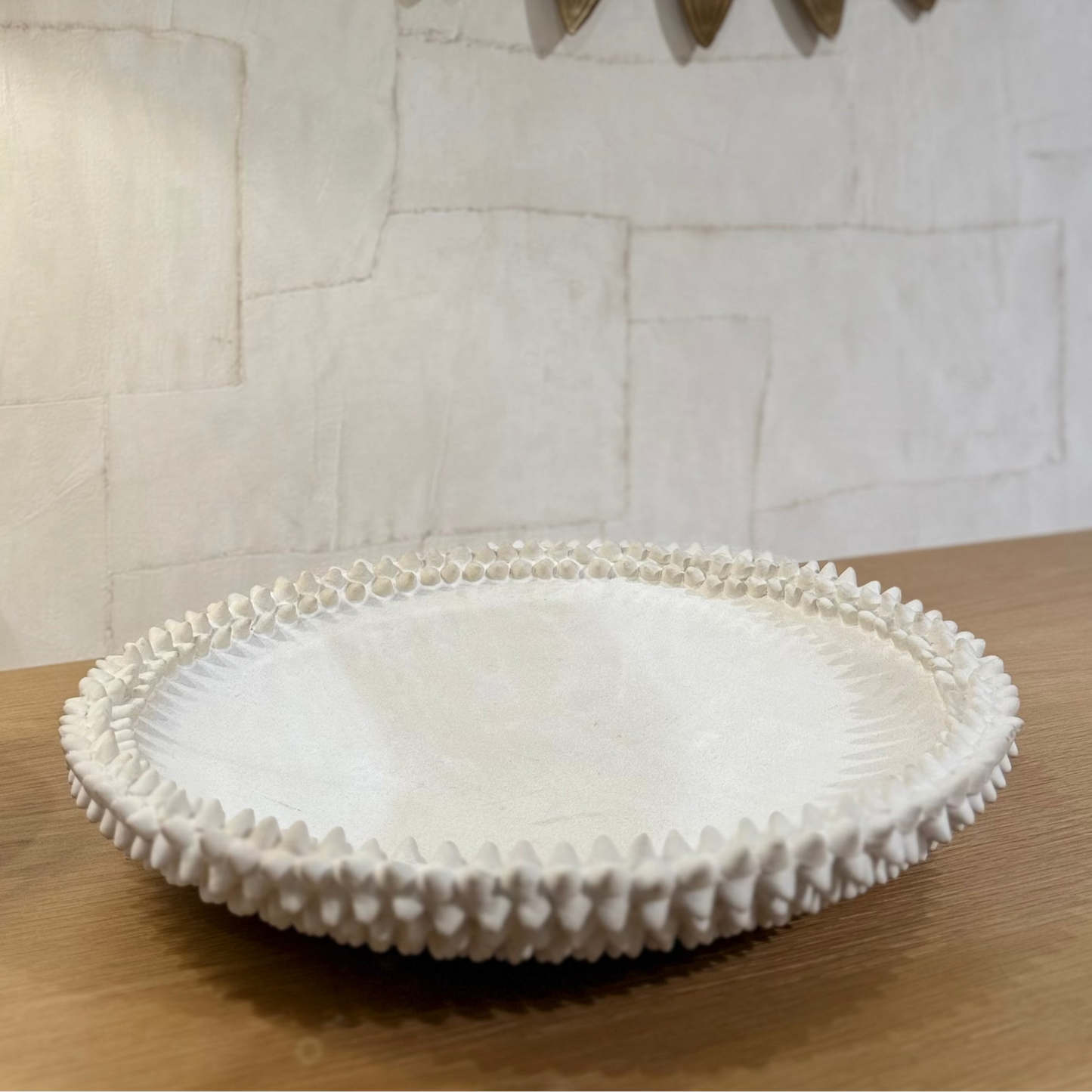 Decorative white dish with textured rim on a wooden surface, with a decorative gold-framed mirror in the background.