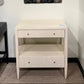 White nightstand with two drawers in a room with mirrors and lamps.