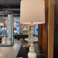 White table lamp with a beige shade on a black surface in a showroom setting.