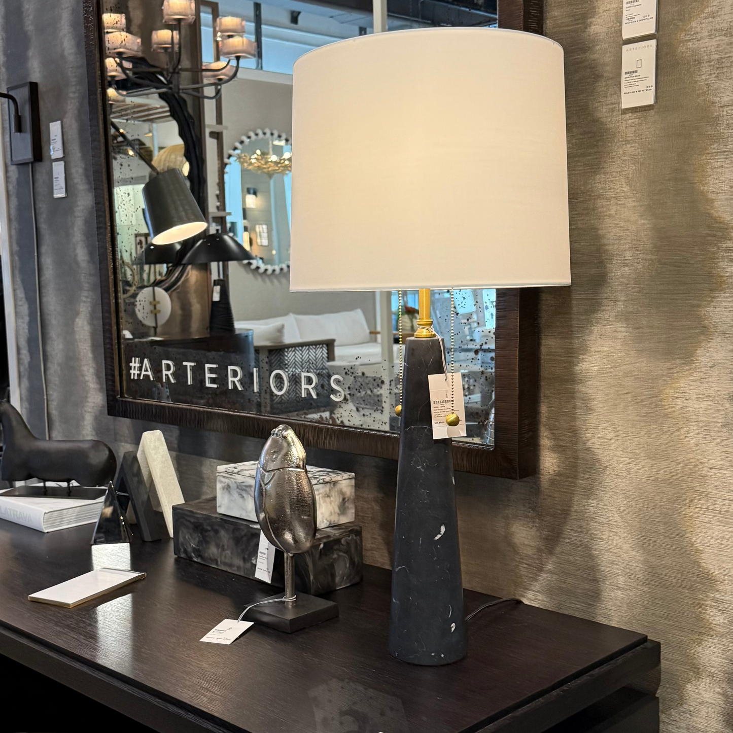 Table lamp with beige shade on a dark surface in a store setting with mirrors and decor.