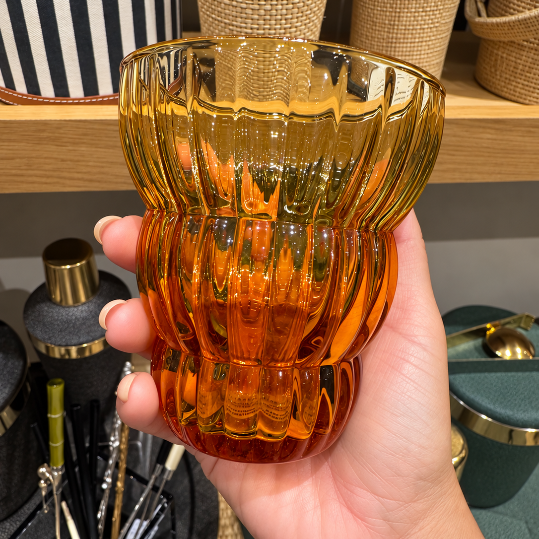 A hand holding a translucent amber glass cup with a ribbed, fluted design, set against a blurred background of home decor items on a wooden shelf.