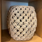 A cylindrical woven pouf or stool cover made from thick, textured cream-colored rope or jute yarn, displayed on a wooden shelf.