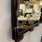 A close-up of a decorative wall mirror with a dark wood frame featuring carved leaf-like corner accents with gold-toned trim, mounted on a white wall in a showroom.