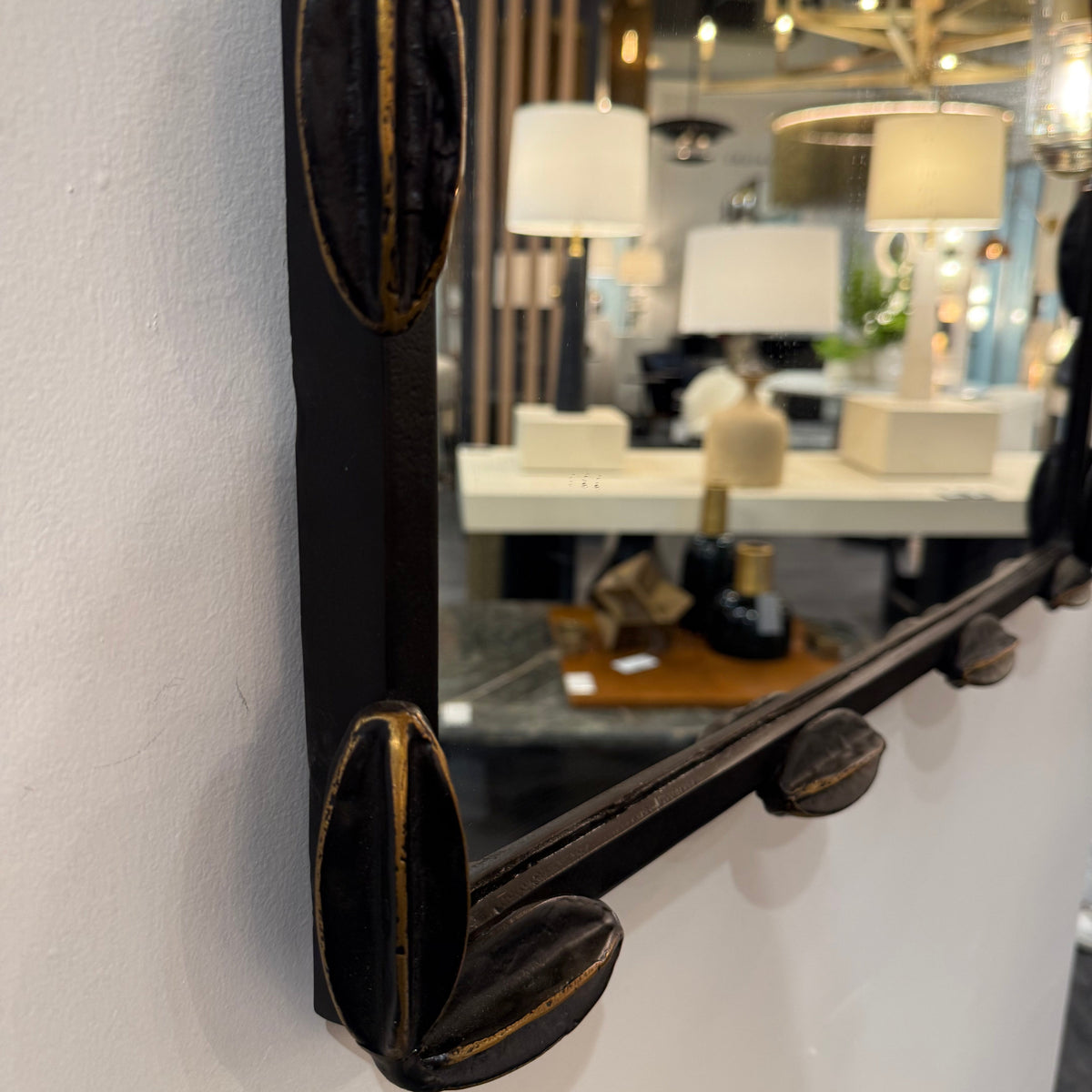 A close-up of a decorative wall mirror with a dark wood frame featuring carved leaf-like corner accents with gold-toned trim, mounted on a white wall in a showroom.