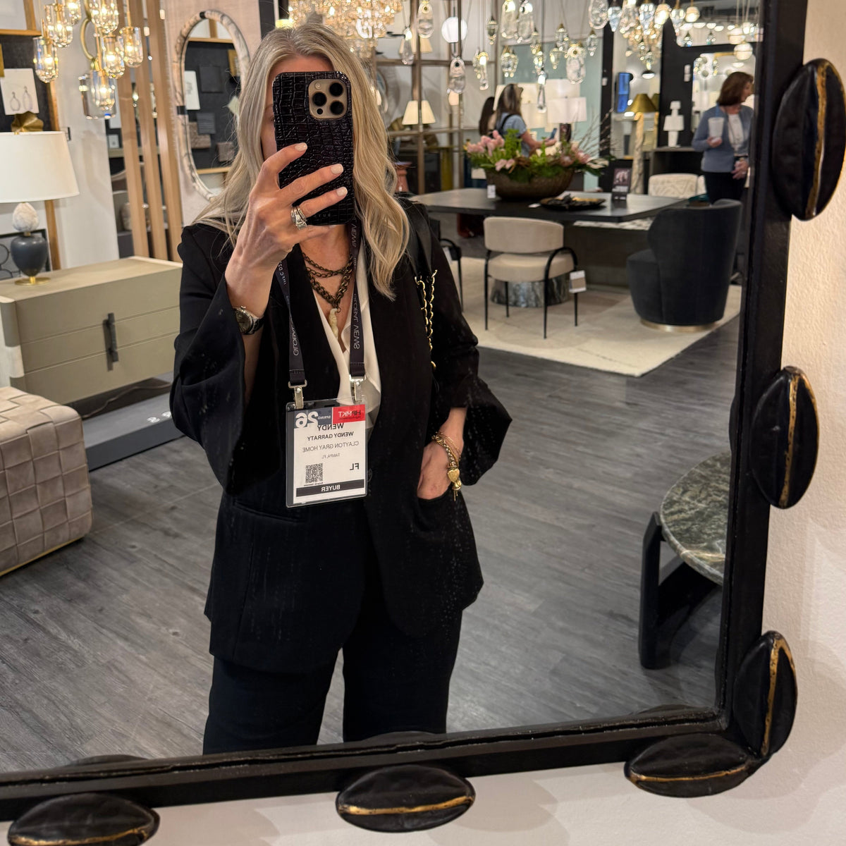 A woman takes a mirror selfie in a furniture showroom, wearing a black blazer and black pants. The background shows various hanging chandeliers, lamps, and furniture pieces in a modern home decor store.
