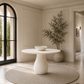 A round white pedestal dining table sits on a large circular woven rug in a bright, elegant room with cream walls, black-framed arched windows, and a tall potted tree.