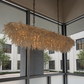 A modern rectangular chandelier with a textured, leaf-like woven shade in a warm natural beige tone, suspended by dark metal chains in a bright interior space with large windows.