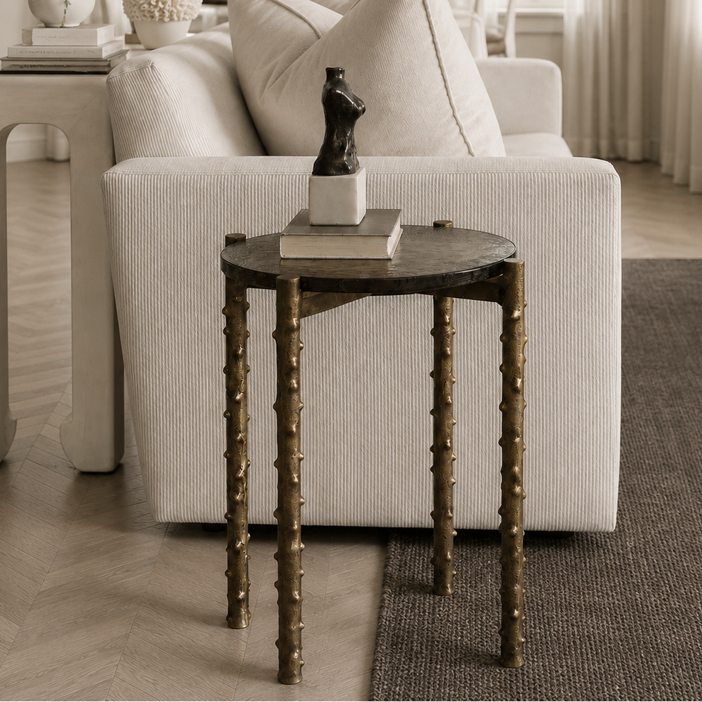 A modern round side table with distressed brass-brown legs and a dark metal top, styled in front of a cream-colored sofa in a bright, neutral-toned living room.