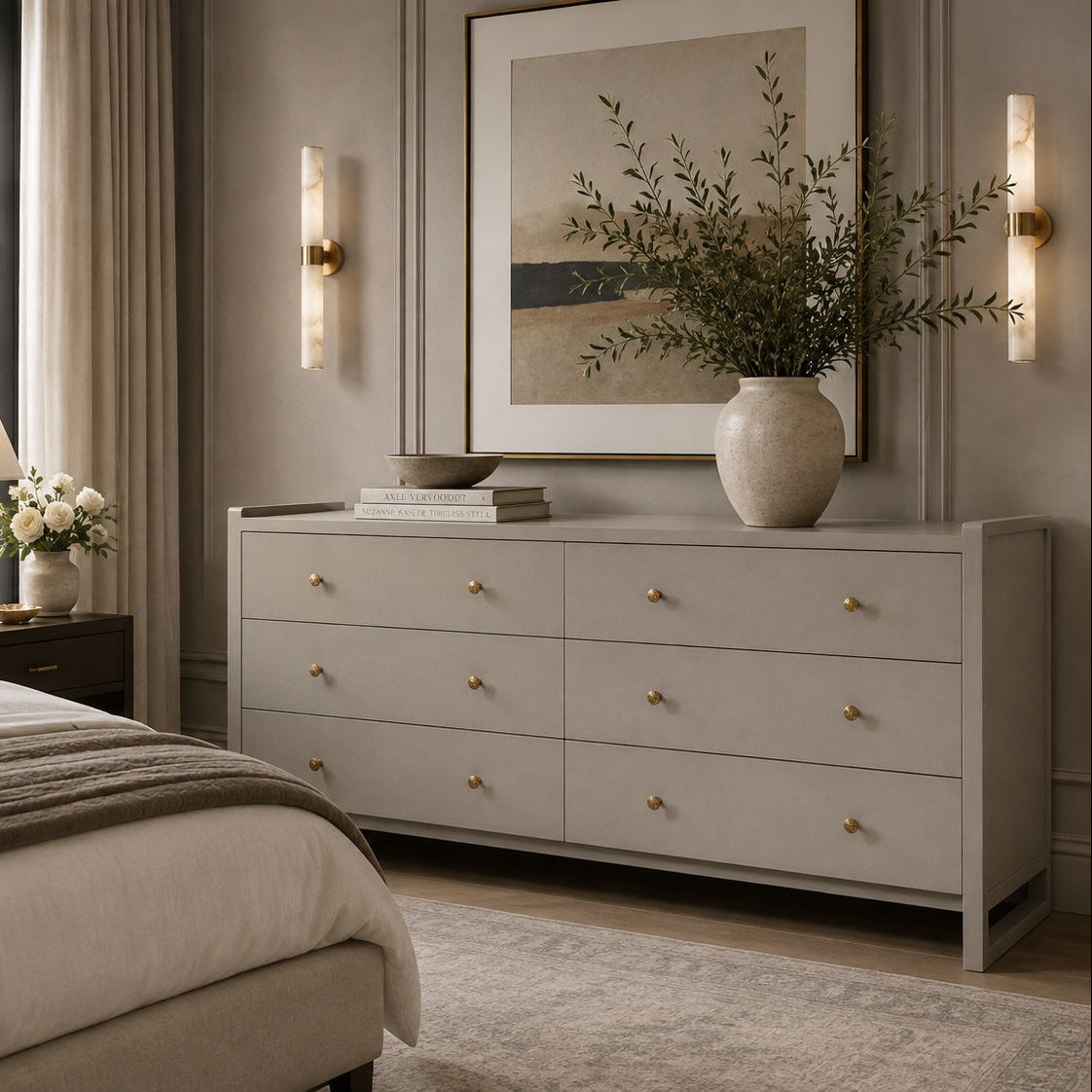 A light gray wood dresser with brass knobs in a styled bedroom setting, decorated with a large ceramic vase of branches, books, and a lamp on a matching nightstand.
