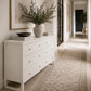 A white six-drawer dresser with small gold knobs in a bright hallway, styled with a large white vase of greenery, books, and a bowl, beneath a framed artwork.