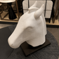 A white sculptural horse head bust displayed on a black rectangular base, set on a dark round table in an indoor showroom environment.