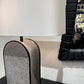 A modern table lamp with a white drum shade and a textured gray arched base with black trim, shown on a dark tabletop next to a black-framed mirror.