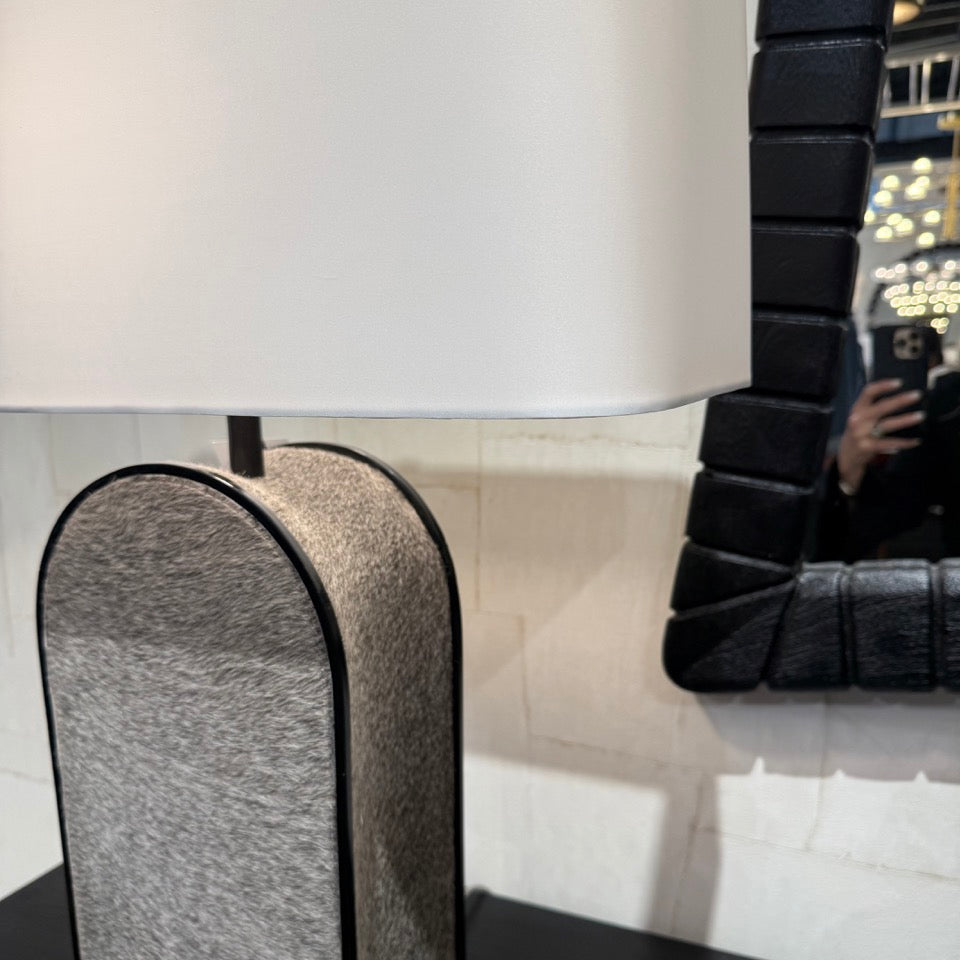 A modern table lamp with a white drum shade and a textured gray arched base with black trim, shown on a dark tabletop next to a black-framed mirror.