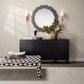 A styled interior scene showing a black sideboard or credenza against a beige wall, with a round gray wood-framed mirror above it, two wall sconces, a vase with greenery, and a black-and-white geometric patterned coffee table in the foreground.