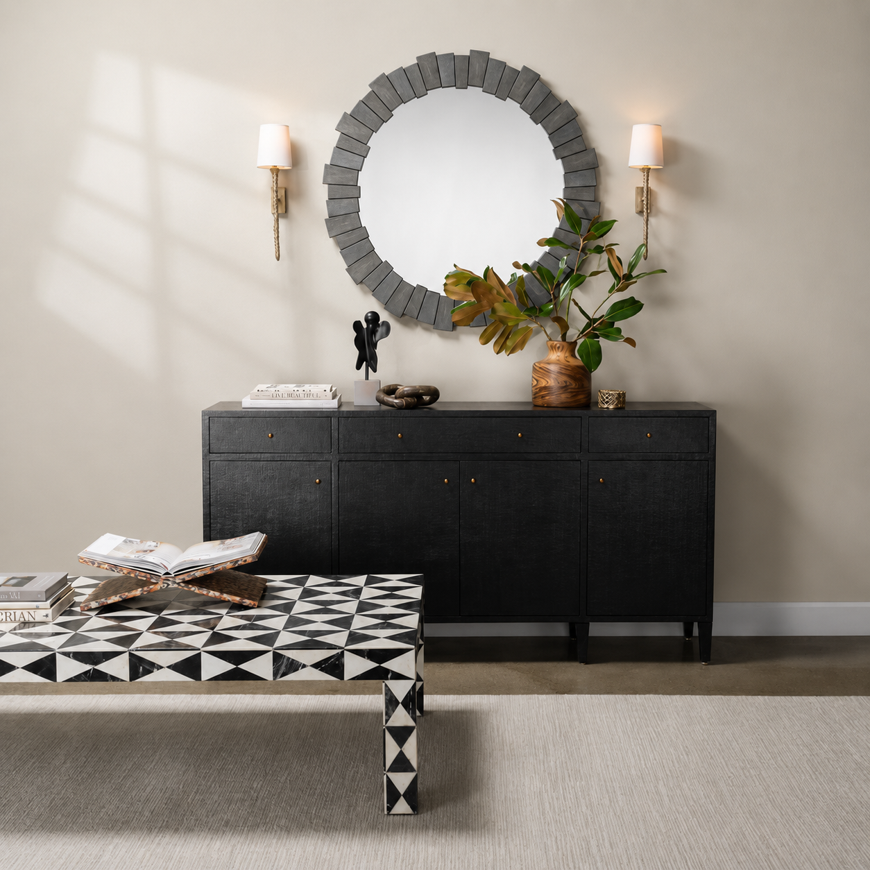 A styled interior scene showing a black sideboard or credenza against a beige wall, with a round gray wood-framed mirror above it, two wall sconces, a vase with greenery, and a black-and-white geometric patterned coffee table in the foreground.