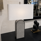 A modern table lamp with a white rectangular drum shade and a textured gray arched base, styled on a black console table in an interior setting.