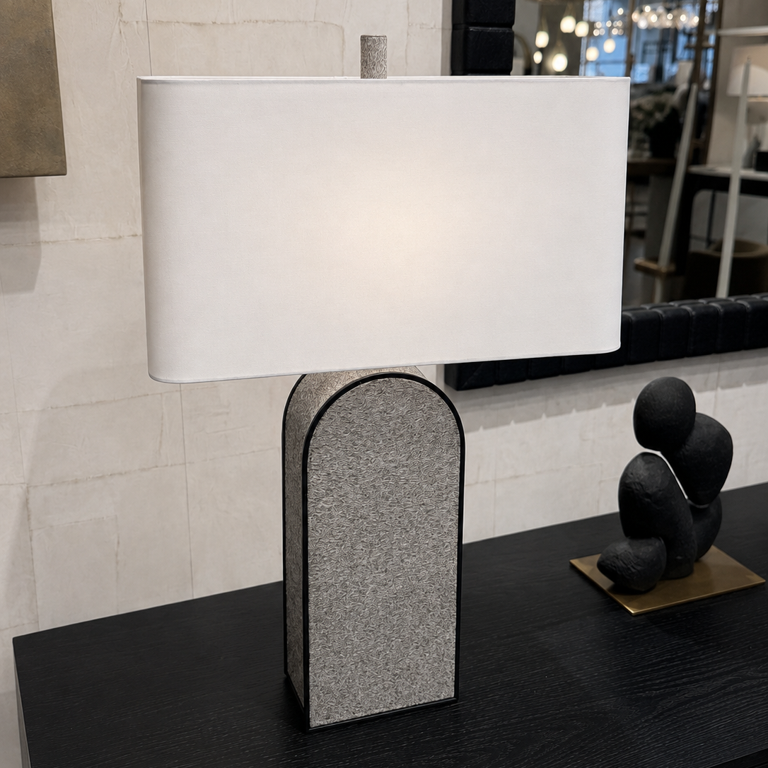 A modern table lamp with a white rectangular drum shade and a textured gray arched base, styled on a black console table in an interior setting.