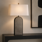 A table lamp with a large white rectangular shade and a textured gray arched base, shown lit on a dark console table against a beige wall.