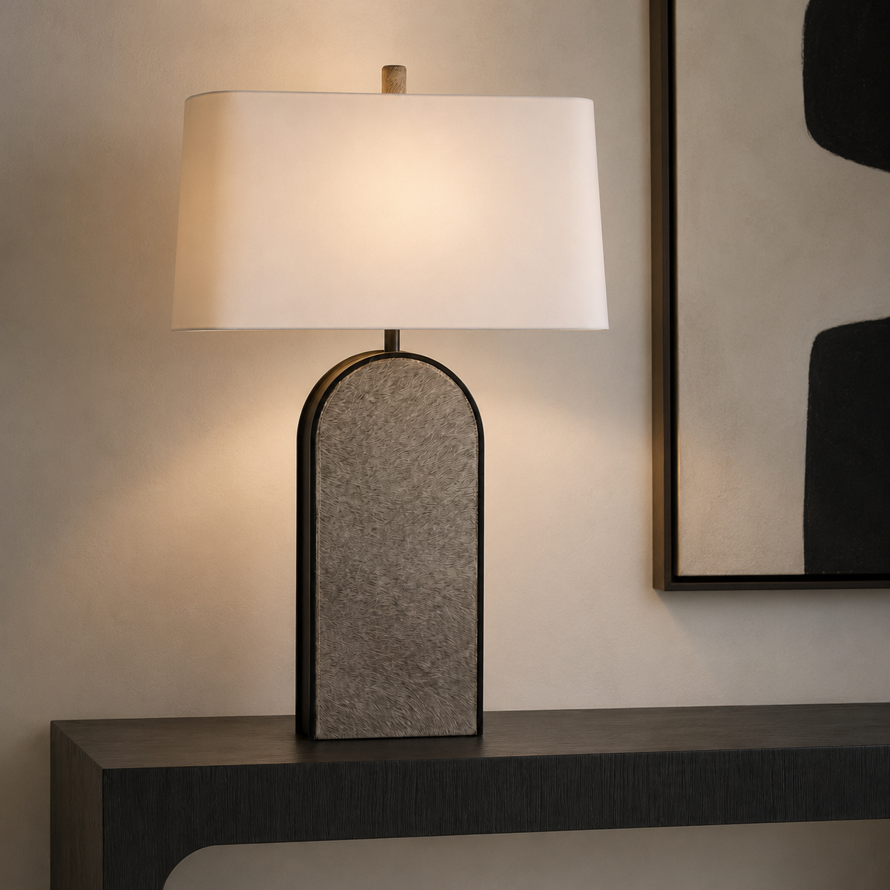 A table lamp with a large white rectangular shade and a textured gray arched base, shown lit on a dark console table against a beige wall.