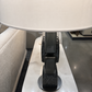 A close-up of a modern table lamp with a large white drum shade and a sculptural dark gray base, set on a white marble side table.