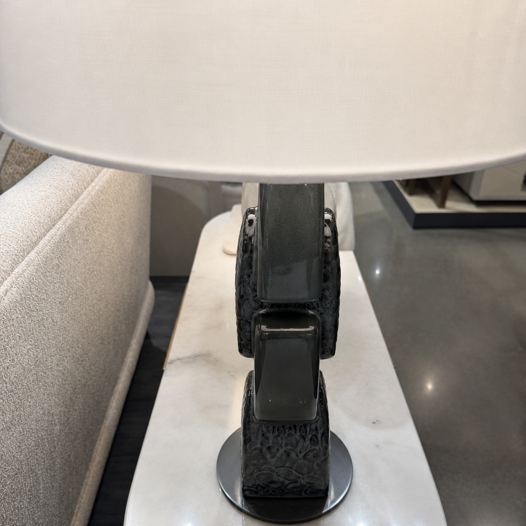 A close-up of a modern table lamp with a large white drum shade and a sculptural dark gray base, set on a white marble side table.