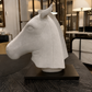 A white sculptural horse head bust displayed on a black rectangular base, set on a dark wood table inside a modern interior showroom with lamps and shelving visible in the background.