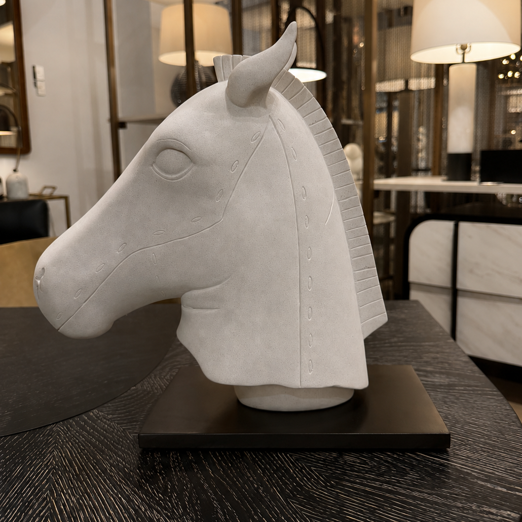 A white sculptural horse head bust displayed on a black rectangular base, set on a dark wood table inside a modern interior showroom with lamps and shelving visible in the background.