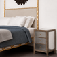 A styled bedroom scene showing a bed with white bedding and a blue throw blanket next to a three-drawer nightstand with distressed gray wood, mirrored panels, and small gold knobs.