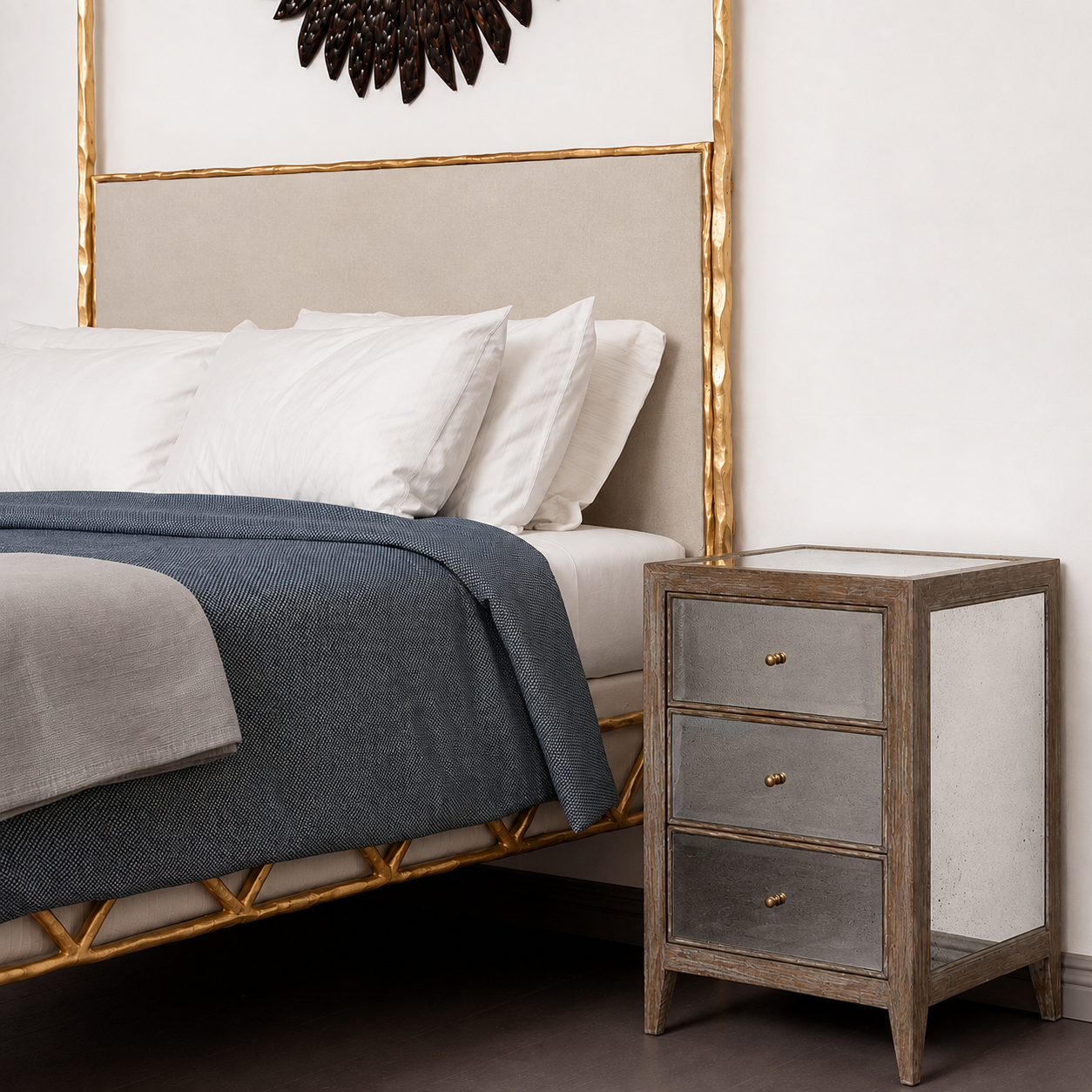 A styled bedroom scene showing a bed with white bedding and a blue throw blanket next to a three-drawer nightstand with distressed gray wood, mirrored panels, and small gold knobs.