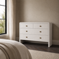 A white six-drawer dresser with small gold-toned square knobs, positioned in a bedroom setting next to a window with beige curtains.