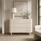 A cream-colored three-drawer dresser with small gold knobs, styled in a bright, modern room with a table lamp and a vase on top.
