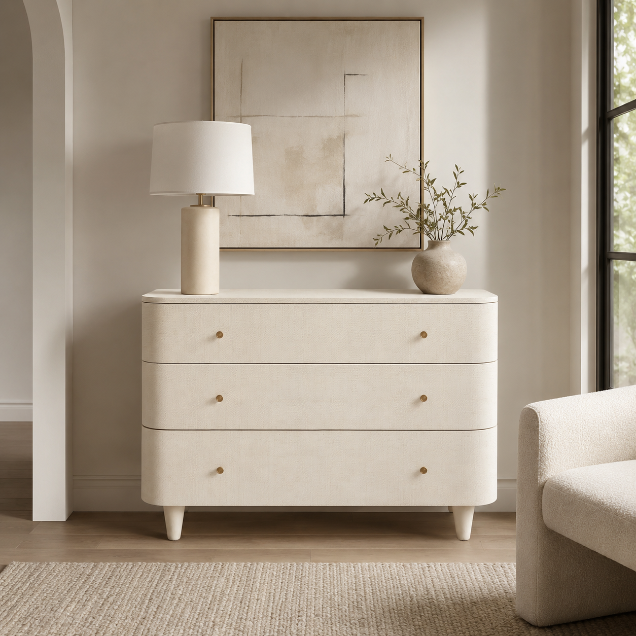 A cream-colored three-drawer dresser with small gold knobs, styled in a bright, modern room with a table lamp and a vase on top.