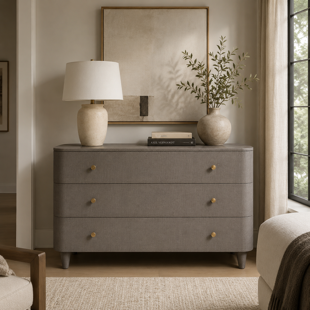 A gray fabric-covered three-drawer dresser with small gold knobs, styled in a warm, neutral-toned room with a table lamp, books, a vase of branches, and wall art.