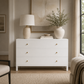 A white three-drawer dresser in a styled bedroom setting, topped with a cream table lamp and a large ceramic vase holding green branches, with small books and a bowl on top.