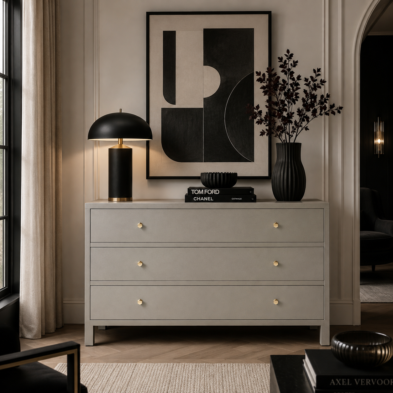 A light gray three-drawer dresser in a styled room setting, decorated with a black table lamp, a black vase with dried branches, and a black bowl. The dresser features small gold knobs and stands against a beige wall with a framed abstract print above it.