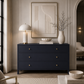 A navy blue three-drawer dresser in a styled living room setting, decorated with a white table lamp, a white ceramic vase with dried branches, a small white bowl, and books.