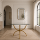A round glass-top dining table with a curved gold metal base, set on a beige geometric rug in a bright, modern dining room with cream walls and wood floors.