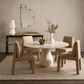 A round dining table with a sculptural pedestal base and four upholstered chairs in a warm neutral dining room setting.